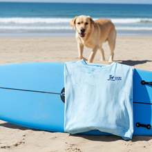 Load image into Gallery viewer, Blue t-shirt with a Lucky Labrador logo on a blue surfboard on a sandy beach with a dog in the background.