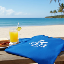 Load image into Gallery viewer, Blue t-shirt with 'Lucky Lab' logo on a beach setting with a glass of juice and sandwich.
