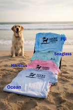 Load image into Gallery viewer, Dog on a sandy beach with Lucky Labrador logo on shirts in various colors.
