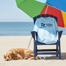 Load image into Gallery viewer, Blue sweatshirt with 'Lucky Labrador Surf Co' design on a beach chair with a dog and colorful umbrellas in the background.