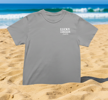 Load image into Gallery viewer, Grey tee shirt with a Lucky Labrador logo on a sandy beach